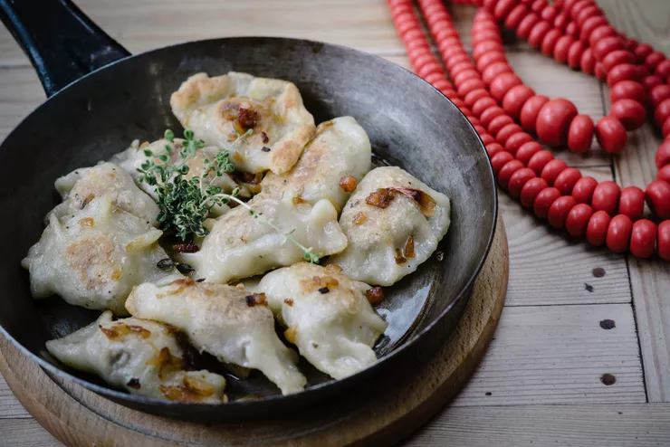 A black frying pan with a serving of nine slightly browned pierogis sprinkled with bits of fried onion is standing on wooden tabletop brushed almost white, next to three strings of large red corals also made of wood.