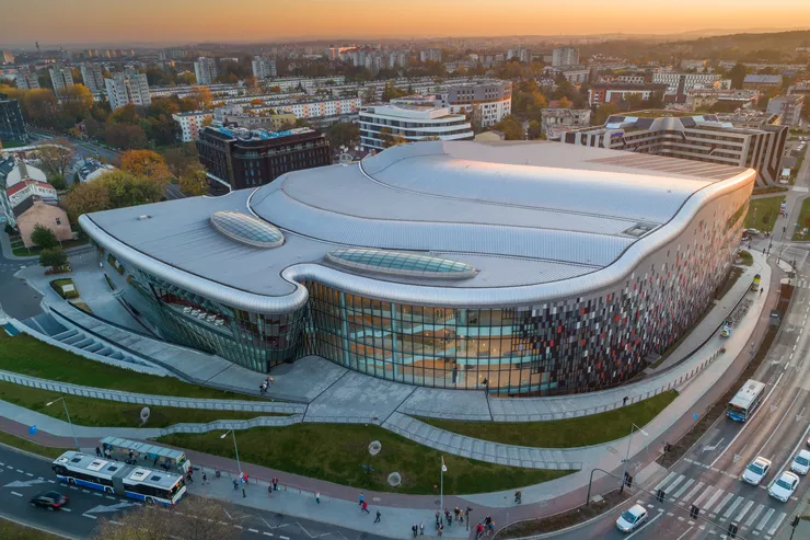 A dusk-time bird’s eye view of the metallic terraced roof and walls of a very large conference building. The wavy façade is fully glazed, and the wall on the right composed of colourful tiles. Two hotels and expanses of residential estates can be seen behind it, and busy streets with people, cars, and busses in front and on the right.