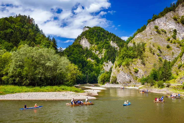 Ein Blick auf den Fluss Dunajec mit mehreren Flößen und Kanus mit Touristen. Rundherum sind zahlreiche bewaldete Hügel mit teilweise herausragenden Felsbrocken zu sehen. Der Himmel ist blau und mit zahlreichen weißen Wolken bedeckt.