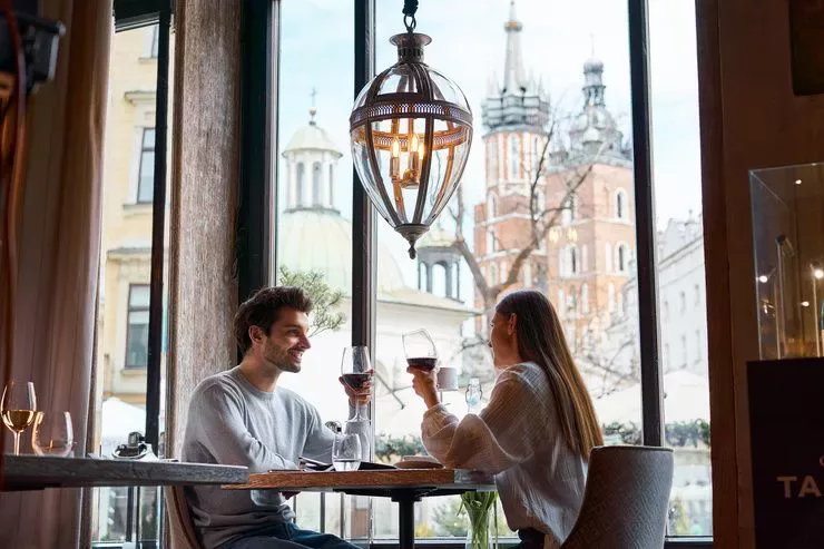 Almost a frog’s eye view at a young man with auburn hair is sitting at a table opposite a blonde woman. The two hold their glasses of red wine in their left hands. The table stands under a large decorative lamp, close to a huge window overlooking sunlit Kraków’s Main Market Square. St Adalbert’s Church can be seen just behind it, with the towers of St Mary’s closing the view.