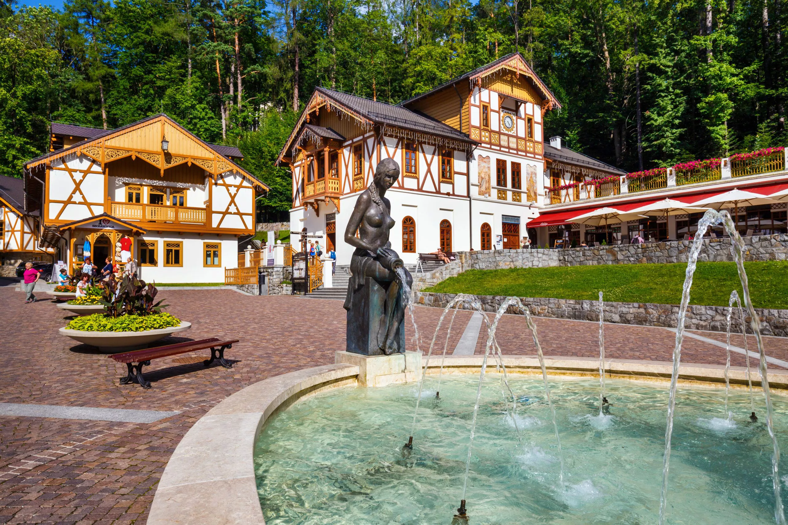 A view of the centre of Szczawnica spa focusing on the Dom nad Zdrojami pump room. There is a round fountain with waterjets and a woman’s statue in the foreground, and Café Helenka above it on the right.