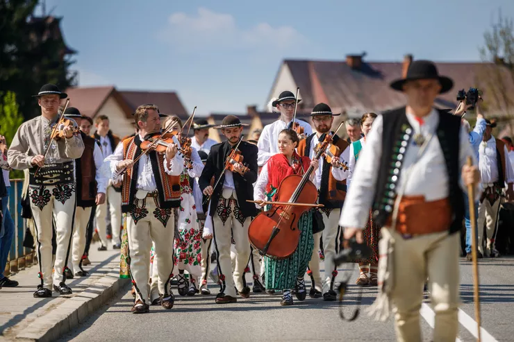 A view of a Highlander band walking along a street. Men and women of various age dressed in regional costumes walk a tarmac road with a strip of pavement on the right. The blurred Highlander in the forgraound is holding a camera in the right and a walking stick in the right hand. Most members of the orchestra are playing the violins, and the woman in the centre wrapped in a red scarf is playing a large cello. The sky over the roofs of the houses in the background is blue.