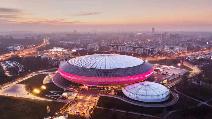 A dusk-time bird’s eye view of a huge strongly flattened sphere of the multifunctional sports and events hall with a magenta strip of lights along its circumference, and grey-and-white dome. It is connected to a smaller hall of a similar shape, font-right. There is a wide artificially lit road leading to the hall, and plenty of residential estates behind it.