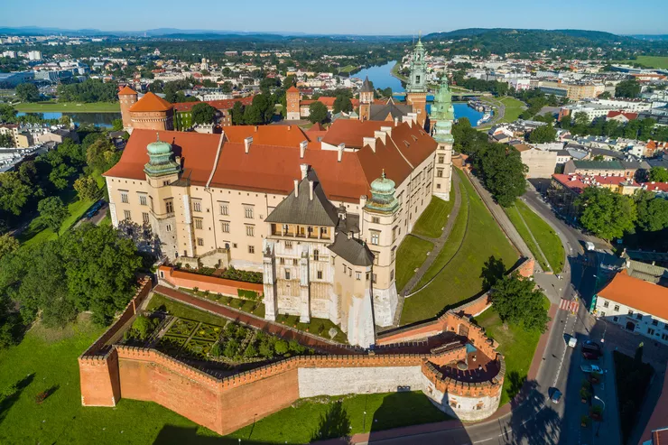 A bird’s eye view of sunlit Wawel Hill, with the ramparts enclosing the Queen’s Gardens in the front, and the walls of the Castle towering over those, slightly dwarfing the Cathedral and towers in the rear. All the tall roofs are made of brick-red tiles, and the spires of green copper sheets. The background consists of clusters of building between which a river is winding its way. The clear sky is blue.