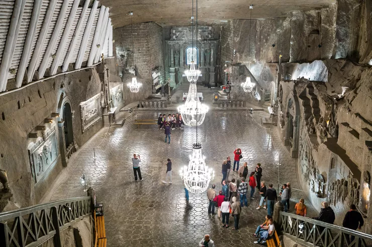 A view of a chamber carved in dark rock salt brightly lit with impressive chandeliers. Standing at the bottom of the stairs leading down to it, and taking the bottom of the photo, are a colourful group of tourists visiting the mine. The far end of the chamber is taken by an altar cut in salt.