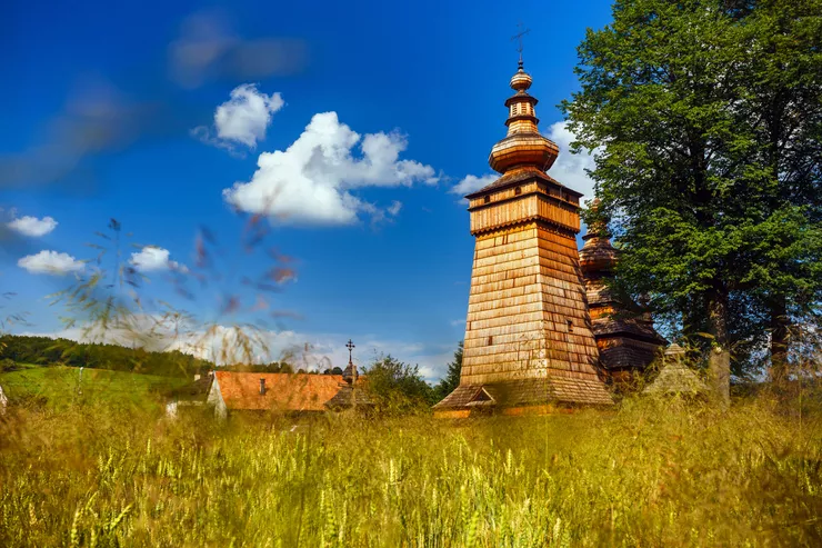 A low shot shows a traditional wooden Eastern church on the Wooden Architecture Route soaring from among the grass growing in the foreground. A cluster of tall trees stands to the right of the church towers, and the there are some tufty clouds in the otherwise blue skies.