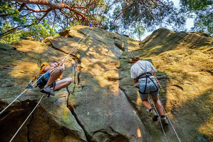 A view of a near-vertical rock face from below, with two people caught resting while top-roping it. Clear blue sky is visible through branches of trees at the top of the rock.