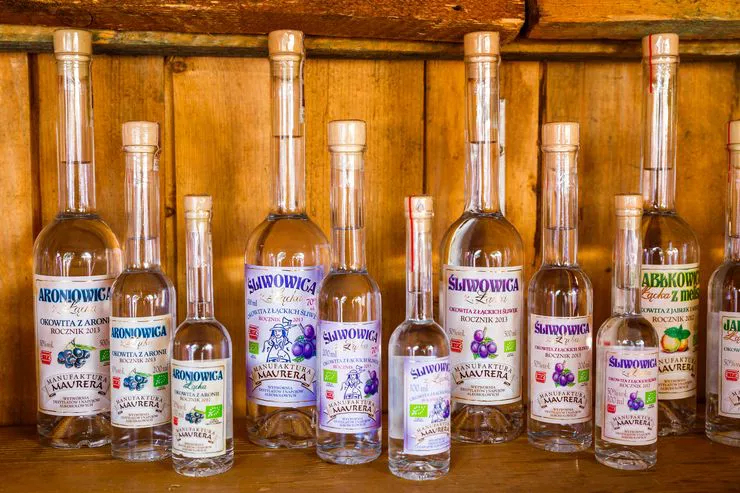 Bottles in three different sizes with different labels, all reading “Śliwowica”, arranged on a wooden shelf against a rustic wall of wooden panels.