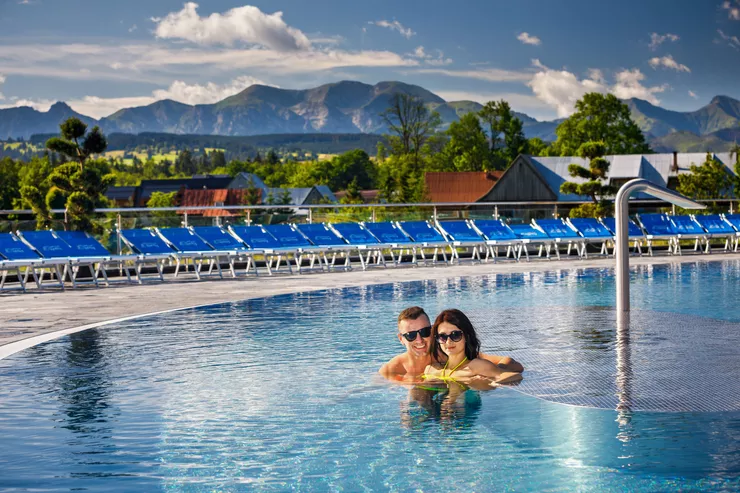 A young couple are taking a relaxing dip in an open thermal pool surrounded by a row of blue deckchairs. The man and woman are immersed in water to the shoulders. A panorama of nearby houses among green trees opens behind them, with the lofty massif of the High Tatras under cloudy sky as the backdrop.
