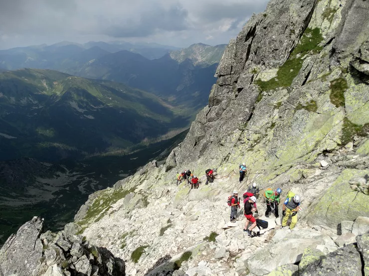 Two groups of tourists in colourful sweaters and helmets are ascending along a rocky ledge on the right, holding to some chains. Unlike the dark ranges of the Tatra Mountains visible behind, the rockface they are climbing is gently lit by the sun.