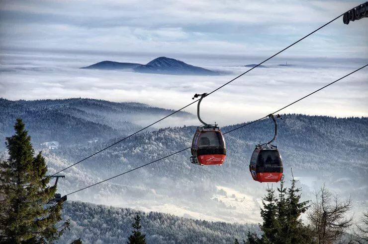 Two red-and-black pods on gondola lift’s cables with treetops below, and a lonely mountain rising from a sea of fog behind snow-swept forested hills.