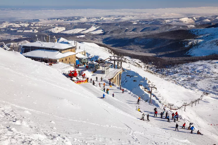 The upper station of the “aerial train” on a snow-clad slope with colourful little figures of skiers on a piste, close to the upper terminal of a ski lift.