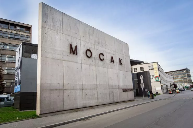 A huge wall made of distinctive concrete slabs bears the five letters making up the name of the museum. A high-rise block is visible behind it on the left, and a long three-storey building further to the right with some cars parked in front.