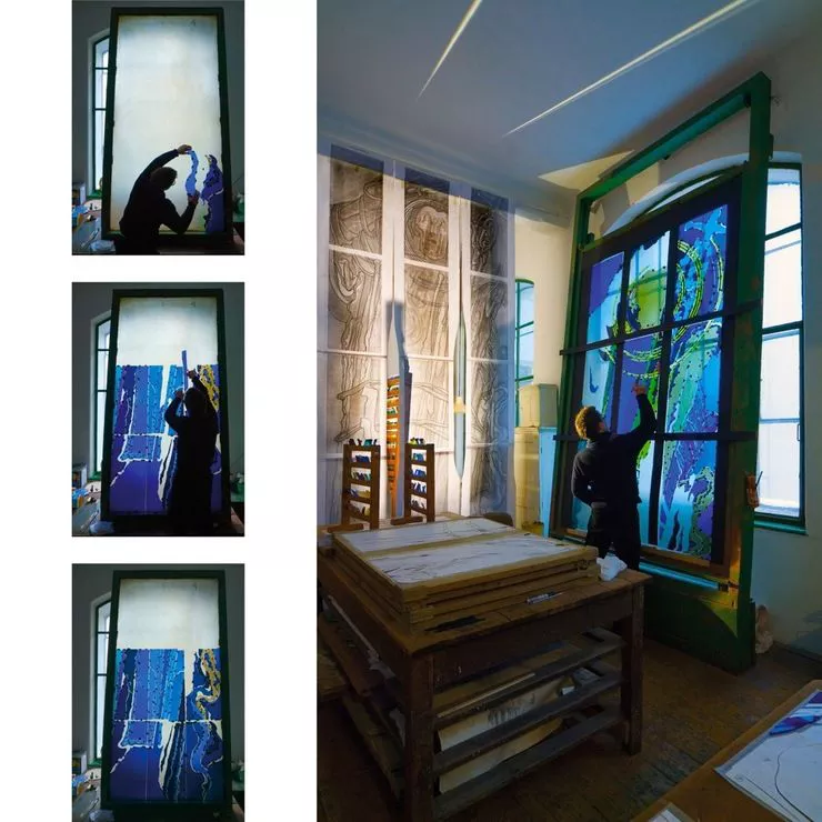 Blick auf einen Mann in einer Glasmalereiwerkstatt, der vor einem großen, bemalten Glasfenster steht, an welchem er gerade arbeitet. An der Seite sind drei kleinere Rahmen zu sehen, auf welchen die aufeinanderfolgenden Phasen der Herstellung der Glasmalerei verfolgt werden können.