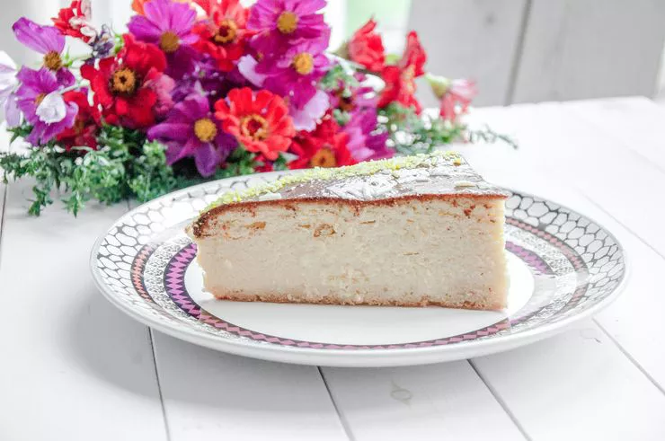 A sumptuous slice of cheesecake on a white plate with colourful border is standing in front of a bouquet of red, violet, and purple flowers with orange centres, on a crude table made of white planks.