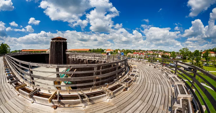 View of the upper terrace of the graduation tower (thorn house) in Wieliczka sided with barriers to protect walkers from falling down from the height onto the surrounding green lawns, behind which white buildings with red roofs can be seen. Standing in the far left against the azure blue sky with some white tufts of clouds is the tallest part of the graduation tower made of countless buckthorn buckthorn twigs and boughs.