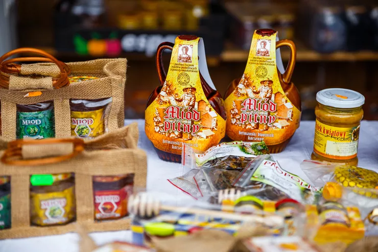 Jars with colourful labels for various types of honey are arranged in special canvas carryalls on the left side of a table, whose centre is taken by an array of apiculture products including packets of bee glue and a jar of pollen lying at the foot of two flagons of mead with labels in the warm colours of honey.