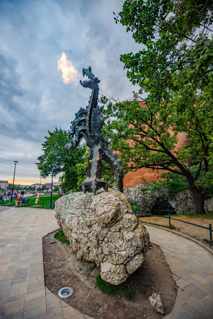 Ein Blick auf die Skulptur des feuerspeienden Waweler Drachens. Die Skulptur des Drachen befindet sich in der Nähe des Wawel-Hügels. Im Hintergrund sind die Drachenhöhle und grüne Bäume zu sehen. Der Himmel ist bewölkt.