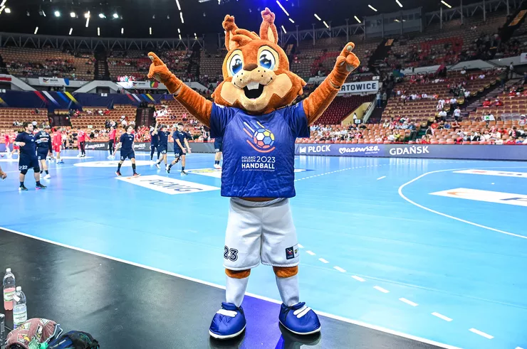 A man with hands lifted into a V is dressed in the costume of the mascot of the World Handball Championships 2023 is standing in an indoor sports arena in front of a blue floor with several players watched from the stands in the background. He is wearing oversized navy-blue shoes, white shorts with a number, an official jersey of the championships, and an even more oversized head of a brown smiling squirrel.