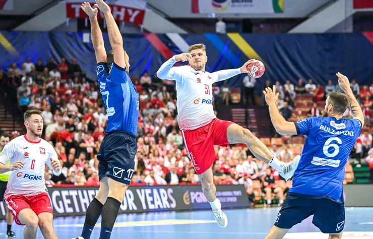 A dynamic photograph showing four men playing handball in an arena with out-of-focus cheering audience and decorations in the background. The two defending players are wearing blue jerseys and black shorts, while the attackers are wearing white jerseys and red shorts. The feet of the attacker in the possession of the ball and the defender blocking him are evidently off the floor.