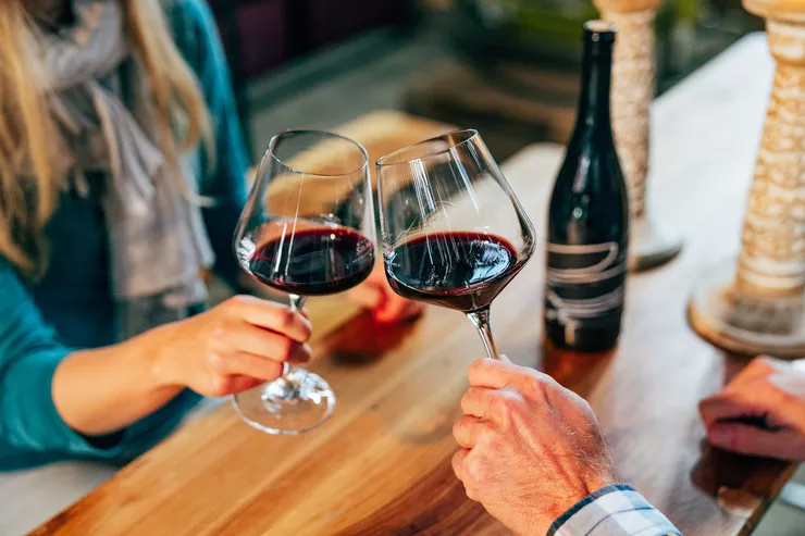 Blick auf zwei Hände, eine weibliche und eine männliche, die Gläser mit Rotwein anheben. Im Hintergrund, auf dem Tisch stehen eine Flasche Wein und hölzerne Gewürzmühlen.