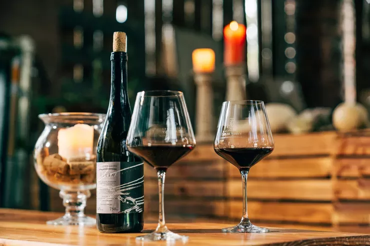 A dark green corked bottle is standing behind two glasses with some red wine on a wooden table that also supports a bowl on a short stem with a candle inside. Two other out-of-focus candles can be seen in the background against some vertical strips.