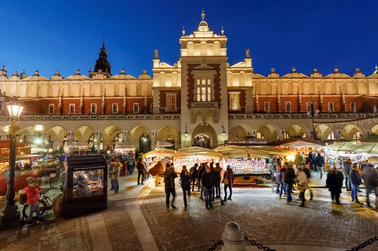 A wide-angle shot of an evening at the Christmas fair in the Main Market Square. Clusters of people are gathered in front of brightly lit stands with assorted Christmas products in front of the warmly lit central section of the Cloth Hall. The top of the Town Hall Tower peers from behind it against a dark blue sky.