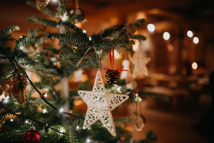 A delicate ornament in the shape of five-pointed star is hanging from a bough of a decorated Christmas tree. Other decorations, including LED lights, are mostly out of focus, visible against the background of a blurred room in subdued brown-auburn light.