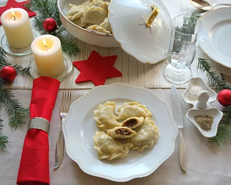 A top view of a wooden table set decoratively for a Christmas feast. Four dough pockets (pierogi), one of them cut into halves, arranged in a large table bottom centre; more pierogi are visible in a tureen standing above it.