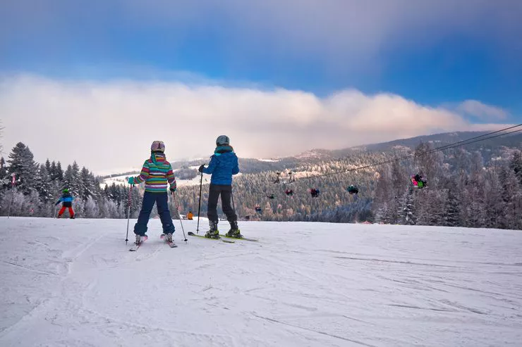 Two young skiers, turned with their backs to us, ski poles in their hands, stand on a gentle crest of a snowy hill looking ahead towards wooded mountains providing a backdrop for a ski lift.