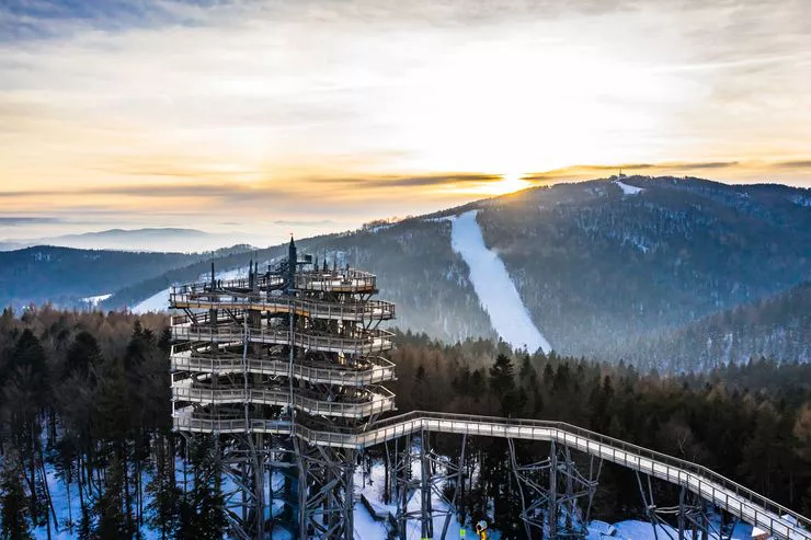 The sun is setting gently over a wooden multi-storey Panoramic Tower that stands on top of the hill overlooking several forested hills, a ski piste cut in the woods on one of them.