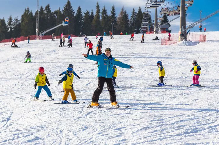 A group of children in yellow vests learn to ski on a gentle snow-covered brow of a hill, following an instructor in a light blue jacket. Several other skiers can be seen behind them under a ski lift and tall spruces partly blocking the clear sky.