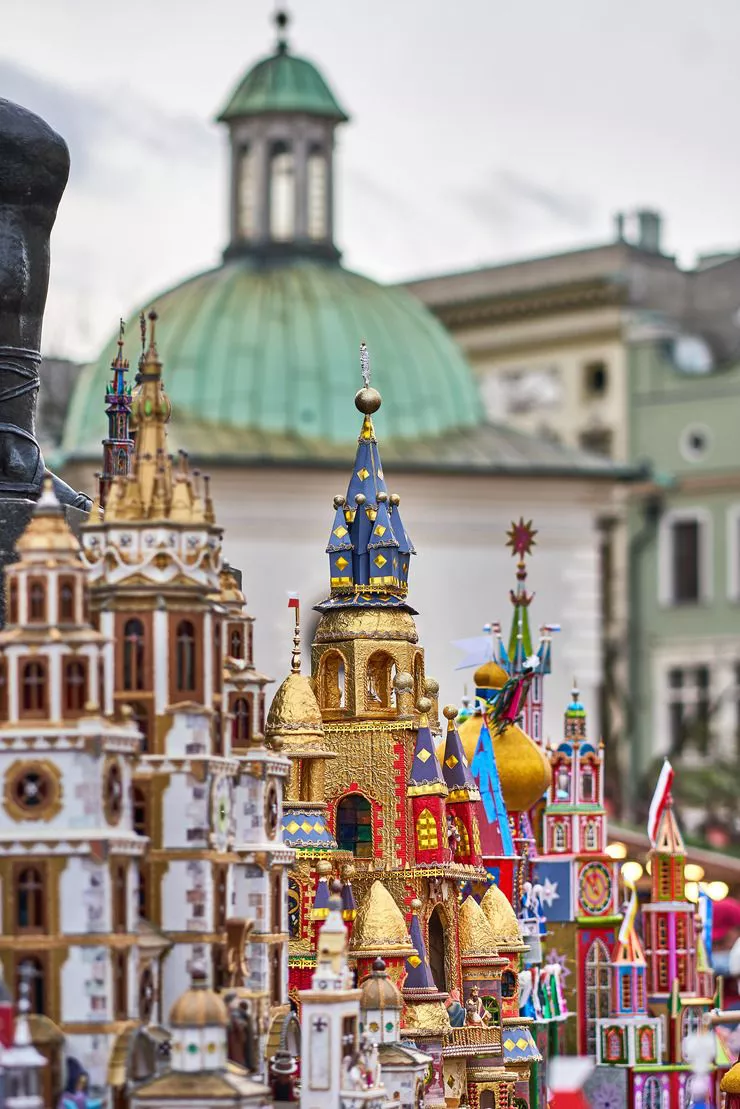 A view of multiple highly colourful multi-spired structures crafted from cardboard and tinfoil displayed against the background of St Adalbert’s Church in the Main Market Square of Kraków.