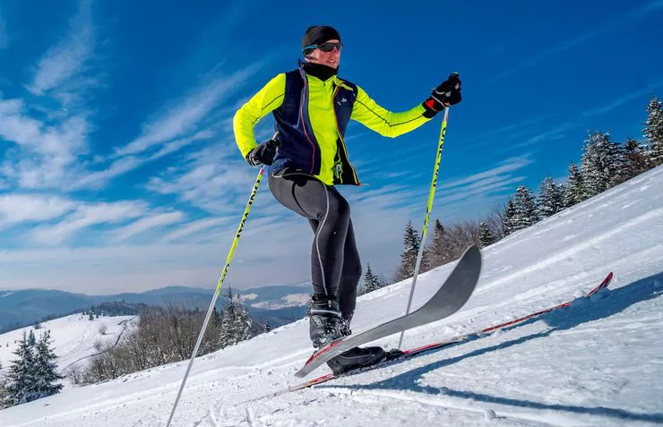 A skier uses two poles to propel himself while cross-country skiing. The hills behind are covered in woods and snow. The sky is blue with tufty of clouds.