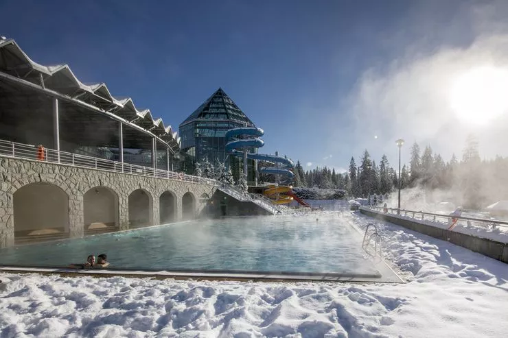A couple relaxing in the steaming spacious outdoor thermal pool surrounded by plenty of snow. The building of Termy Bukovina stands on the far left and sports a twisting waterslide. The sky above is blue.