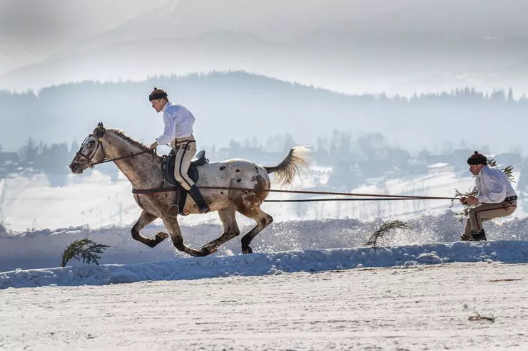 A team of two highlanders participate in a ski-skiring racing on the snow. One of them, the skier, is towed by the other, riding a horse. Both the contestants are wearing traditional Highlander costumes. Many mountain ranges can indistinctly be seen in the background.