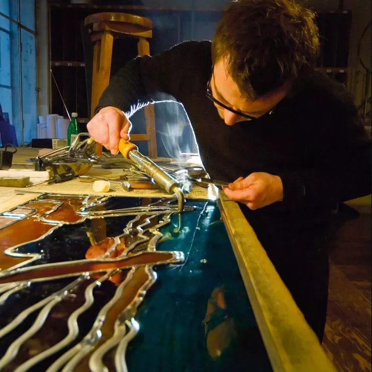 A young man is leaning over a wooden table while working on a colourful stained-glass decoration. The furnished studio where stained glass is made provides the background.