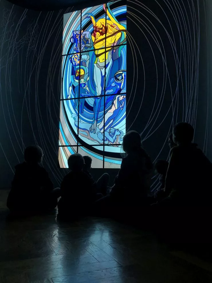 A view of a group of young children sitting in darkness on a wooden floor, admiring a colourful stained-glass decoration strongly lit from behind.