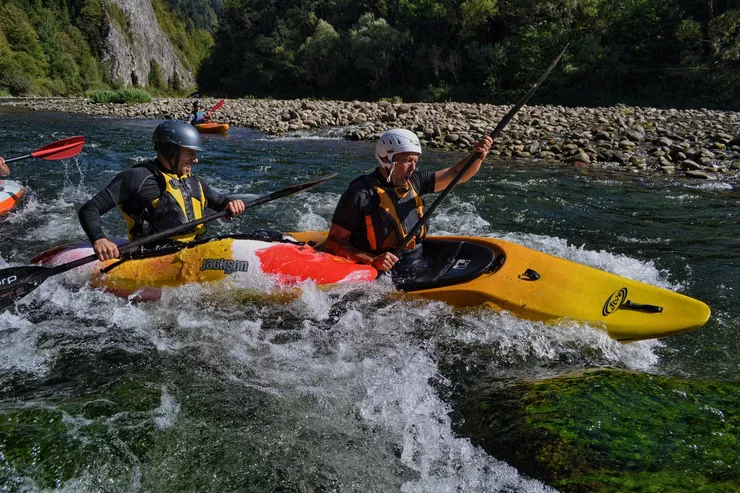 Two people in two canoes go downstream among the white waters of a mountain river. They are both wearing crash helmets and safety vests, and holding paddles. The pebbly shore in the background ends in a wooded hill and a rockface in the distance.