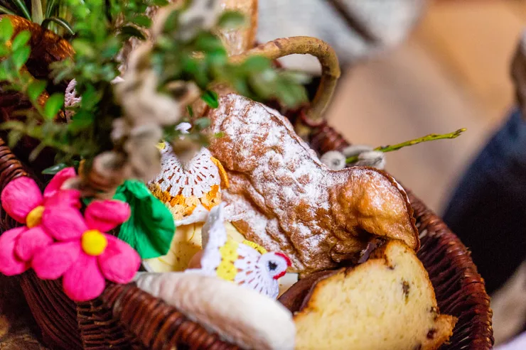 A traditional wicker Easter basket containing among others a bread baked into the form of a lamb, cottage cheese, and Easter decorations. The basket itself is decorated with boxwood and colourful paper flowers.