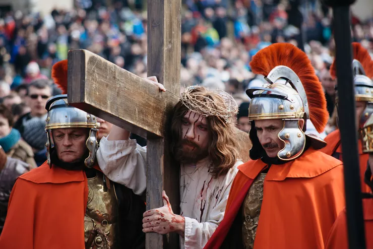 A scene from the Biblical drama showing Jesus in a crown of thorns carrying a wooden cross among red-robed Roman guards wearing crested helmets. Every year, the monks from the local Bernadine monastery and actors play the biblical roles. The background is an out-of-focus crowd witnessing the mystery play.