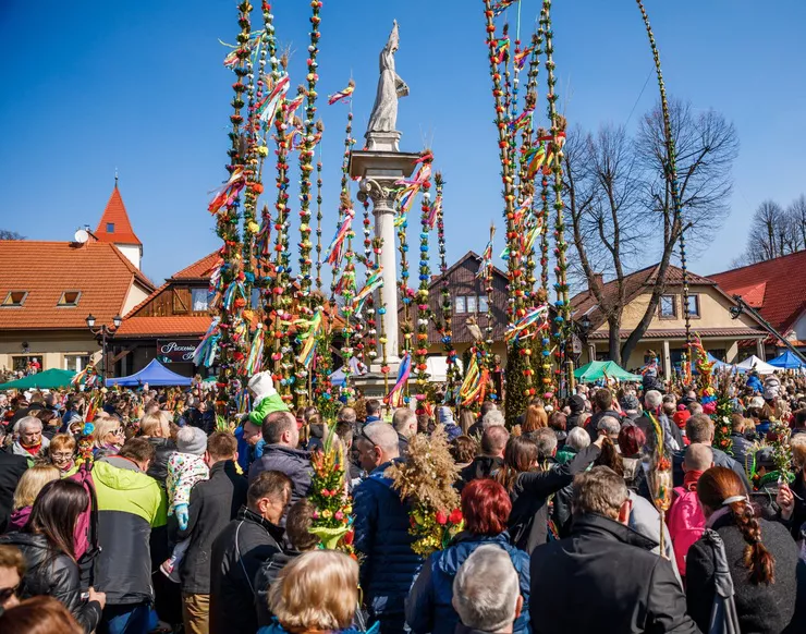 A view of the figure of St Simon on a column surrounded by beautiful Easter palms, very tall and colourful, submitted to the annual competition. The palms are made of willow reeds, and decorated with boxwood, and colourful flowers and ribbons, mostly made of tissue paper. The figure is surrounded by a crowd of people admiring the beautiful palms under the blue sky against the tall roofs of the market square.