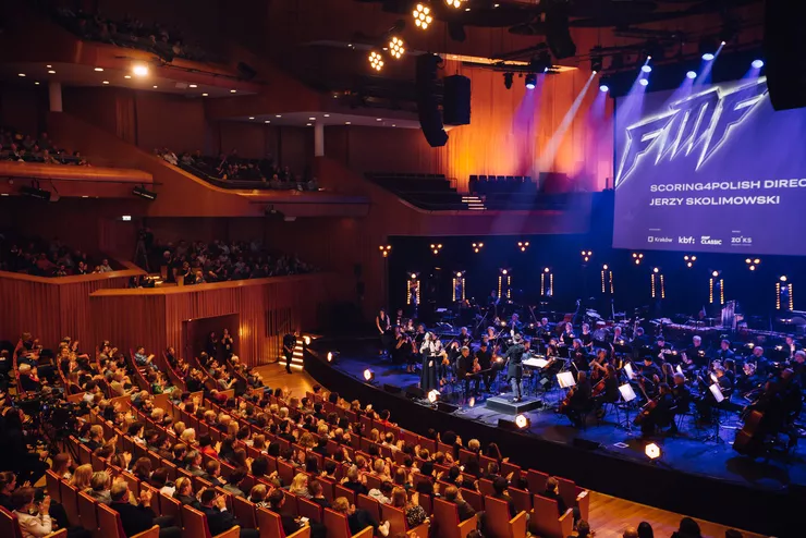 Inside the Main Auditorium of the ICE Kraków Congress Centre, an orchestra perform on the stage under a big projection screen sporting the Kraków Film Music Festival logo. The hall is full, with all the seats in the stalls and boxes taken.