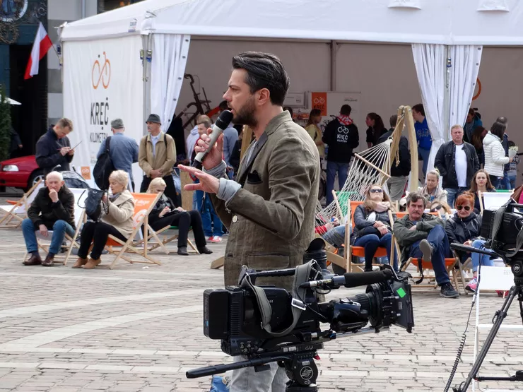 A casually dressed young man is standing behind a camera on a low tripod and holding a cordless microphone. The background consists of a white tent set up in a public square and groups of people sitting in deckchairs in front of it.