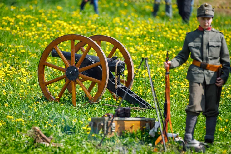 A boy wearing a khaki First World War uniform is holding an old rifle in his right hand. Left from him, in the centre of the picture, there is a small cannon with large wooden wheels standing on a meadow with yellow flowers.