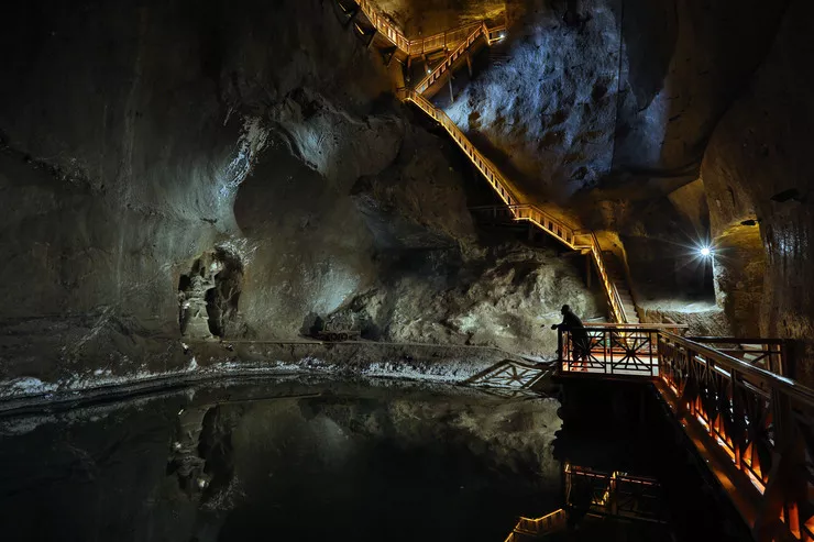 A view of a beautifully though dimly lit Weimar Chamber with a small brine lake at the bottom. A panoramic gallery on the right hand side continues into stairs running up the salt wall in the background. There is a salt sculpture standing by the lake.