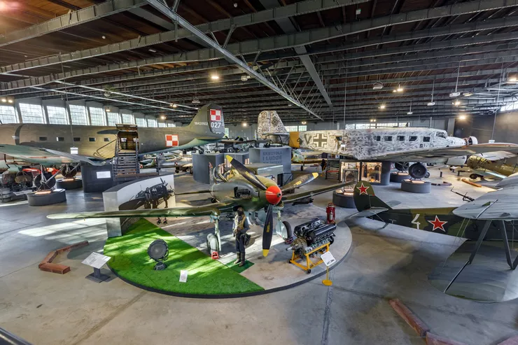 Artificially lit hangar in the museum filled with plenty larger and smaller aircraft. Some of them are fighter planes.