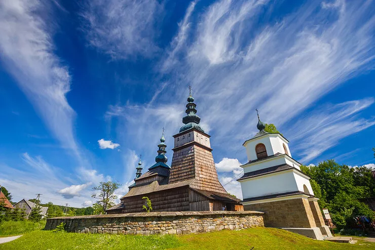Ein Blick auf die orthodoxe Lemken-Kirche, die auf der Route der Holzarchitektur in Małopolska (Kleinpolen) liegt. Es handelt sich um eine dreiteilige Holzkirche mit einem Turm aus einer Pfosten-Rahmen-Konstruktion über der Apsis. Die Dächer über den drei Teilen der Kirche sind mit Schindeln gedeckt. Das Kirchengelände ist von einer Steinmauer mit zwei Eingängen umgeben. Der blaue Himmel ist mit weißen Wolken bedeckt.