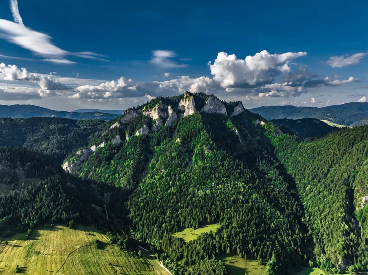 Ein Blick auf den bewaldeten Bergzug Pieniny mit dem Berg Drei Kronen und sonnige Wiesen und Felder. Der Himmel ist blau und mit einigen wenigen weißen Wolkenfetzen bedeckt.