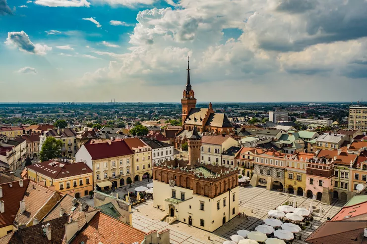 A bird’s eye view of Tarnów Market Square with its central Renaissance townhall with a tower and townhouses surrounding it. Many café and restaurant tables are standing under large umbrellas in the square. The background is made up of multiple buildings, their skyline dominated by an impressive church with a tall spire visible against an overcast sky.